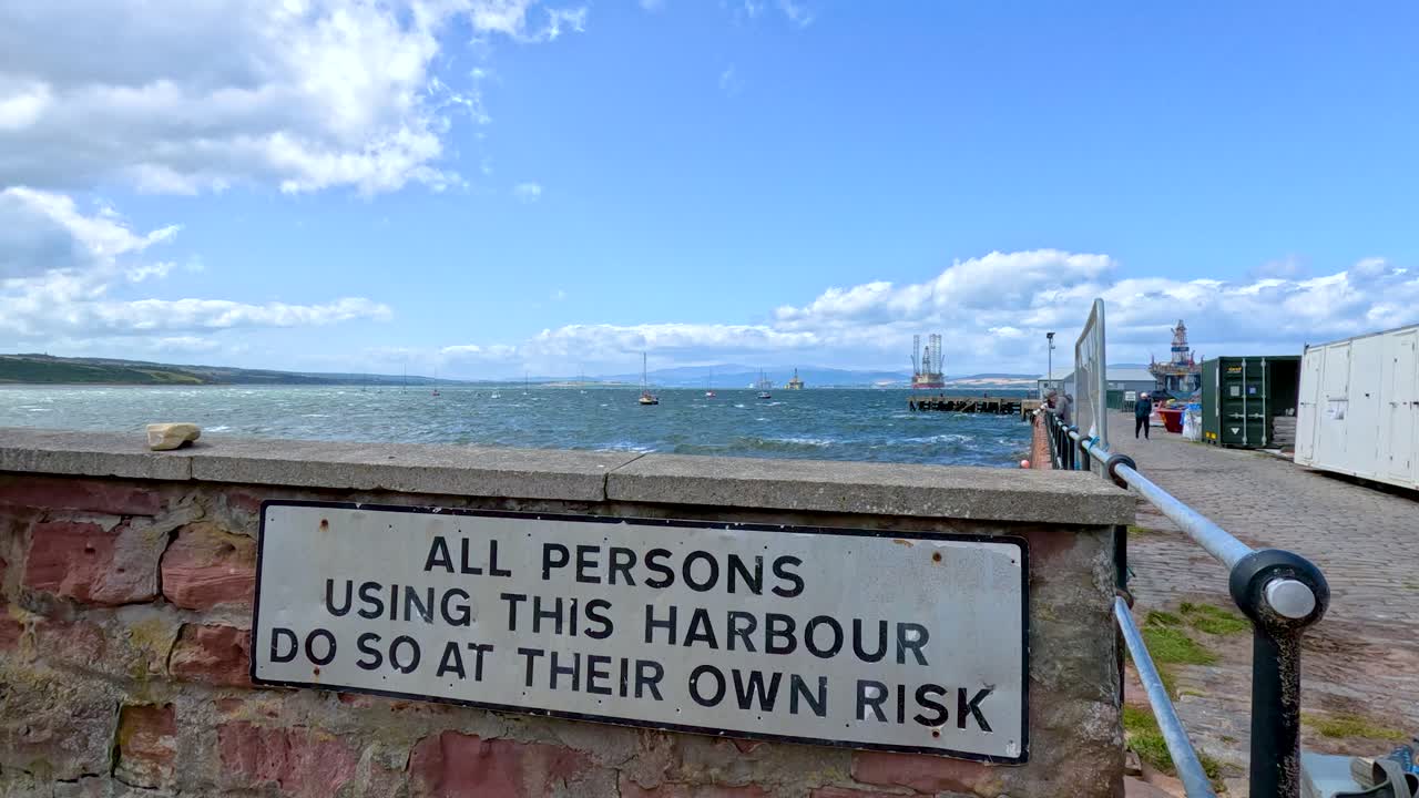 A camera pans across a harbour wall with a safety warning sign, overlooking the sea under bright daylight and scattered clouds