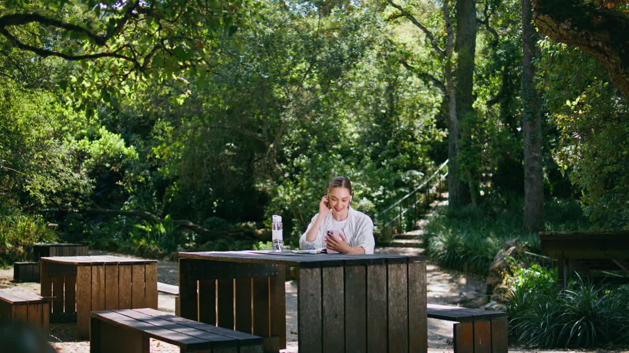 아름다운 여성이 스마트폰 카메라를 인사하고 녹색 공원에서 나무 테이블에 앉아 있습니다.
