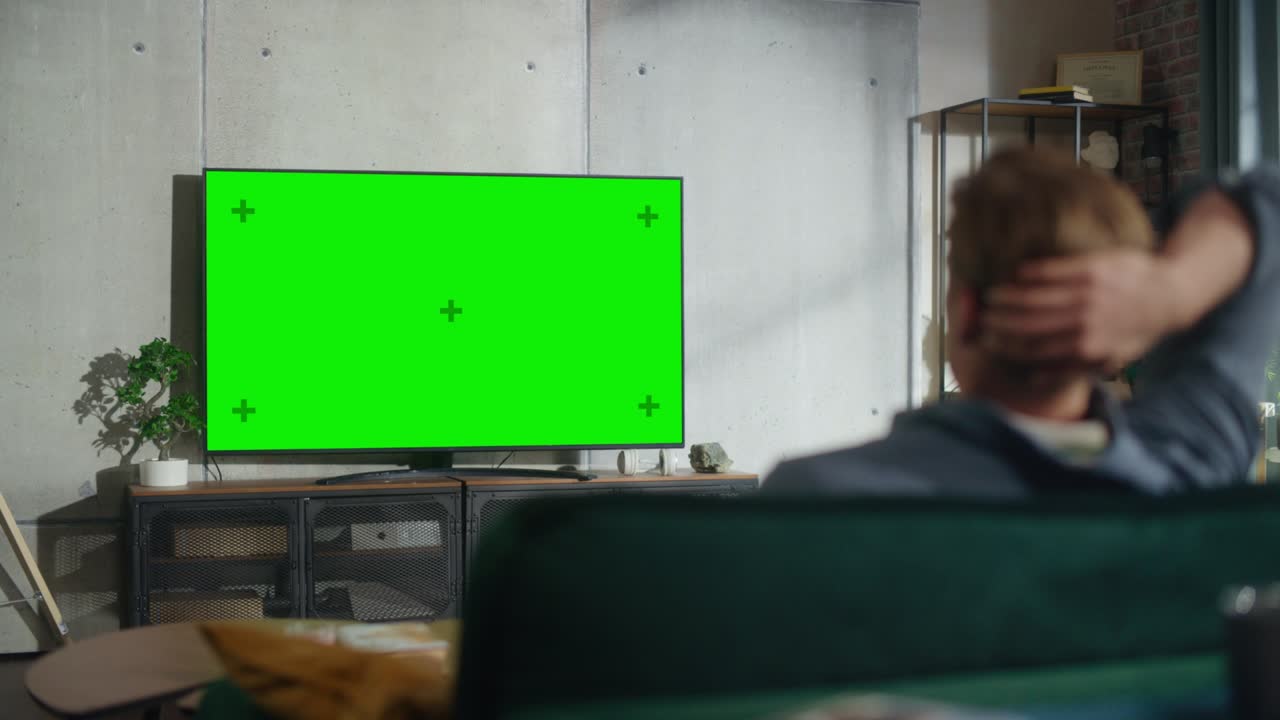 소파에 앉아서 수평 녹색 화면으로 tv를 보고 있는 젊은 멋진 남자, 그것은 집에서 방에서 낮 시간입니다.