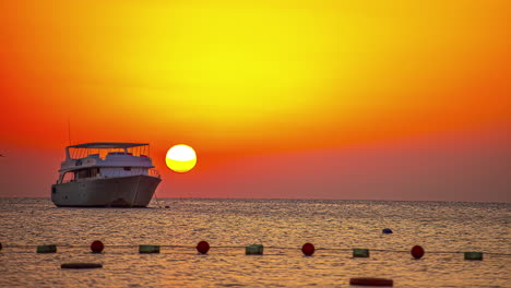 luxury boat anchored on ocean water in front of orange sunset time in background - time lapse