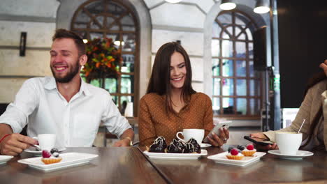 a group of men and women in office clothes sitting in a cafe at a large table and show each other in mobile phones funny pictures