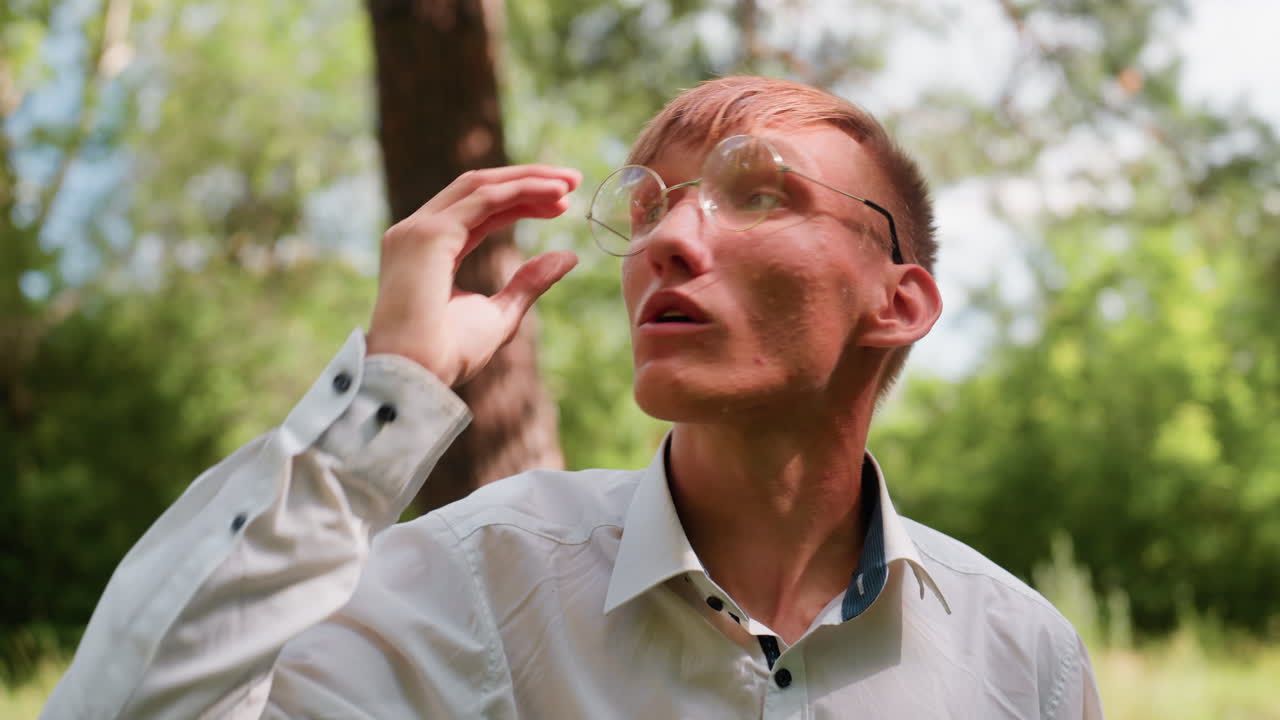 Close view of young man in white shirt raising glasses while whistling with mouth and observing trees in natural forest setting, surrounded by green foliage, expressing attention