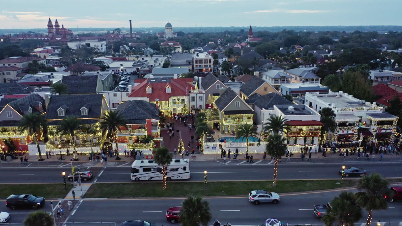 Historic downtown street scene featuring decorated buildings with holiday lights, palm trees wrapped in lights, busy traffic on the road, and pedestrians gathering during dusk hours.