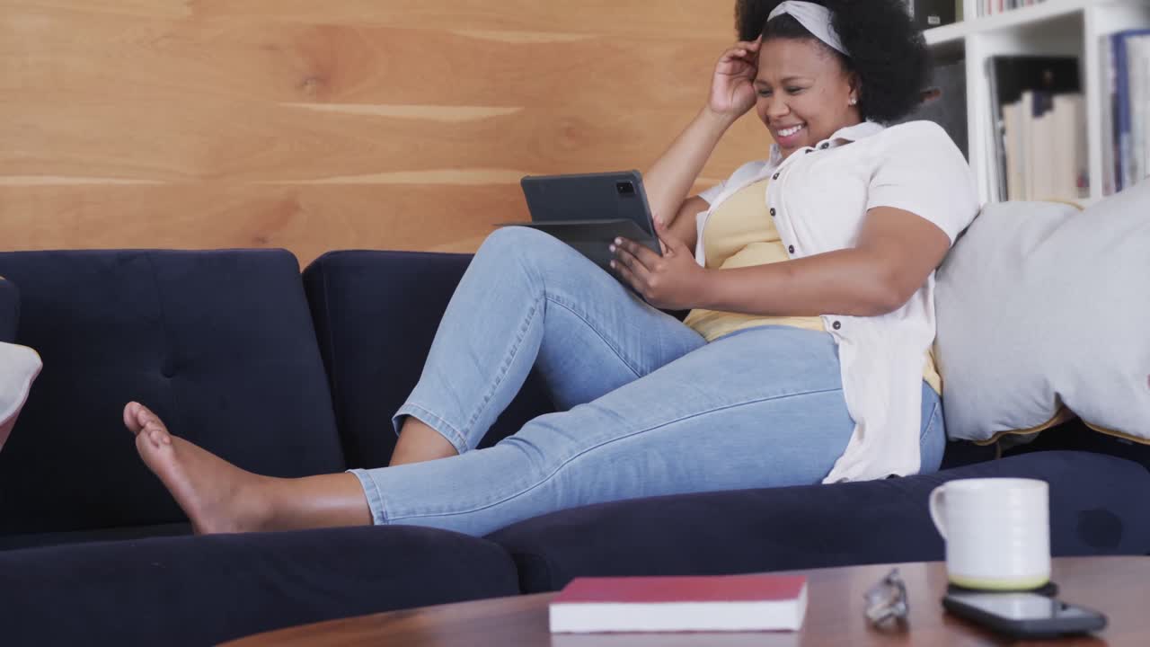 Happy african american plus size woman sitting on sofa and using tablet, unaltered, in slow motion