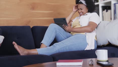 Happy african american plus size woman sitting on sofa and using tablet, unaltered, in slow motion