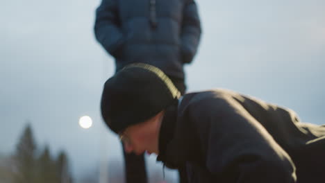 close-up of a young boy in a black outfit and glasses performing push-ups outdoors, with another person standing nearby, with soft light reflecting on him