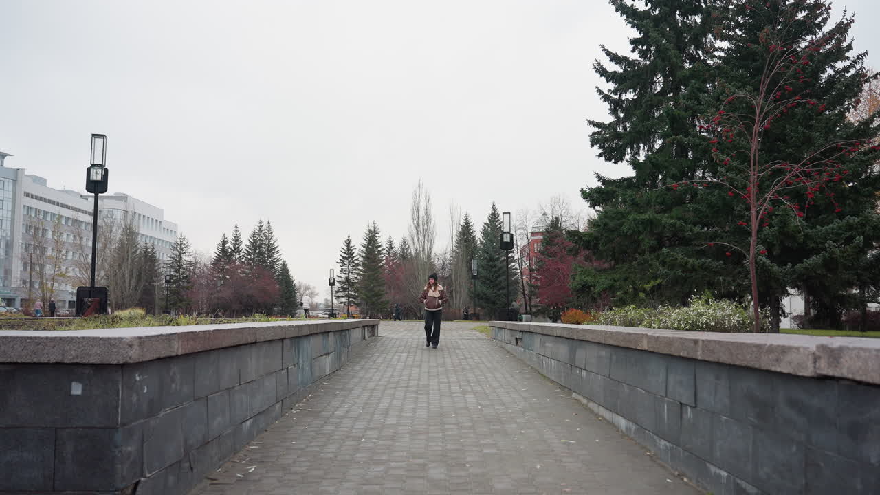 Lady wearing black cap brown jacket and trouser walking confidently on urban park pathway hands in pockets surrounded by trees buildings and cloudy sky in calm autumn city setting