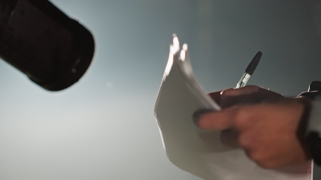 Hands flipping notes near microphone, astronomer preparing observation log inside dim observatory, pen marking pages under soft spotlight, closeup of fingers turning sheet, ambient glow and focused