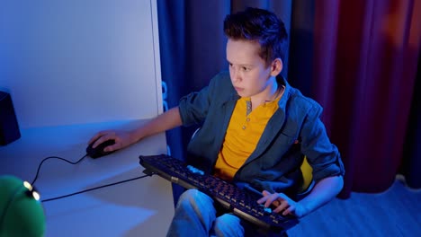gamer playing games on a pc at home. boy is pushing buttons on a keyboard on the desk while playing games in his room. blue light.