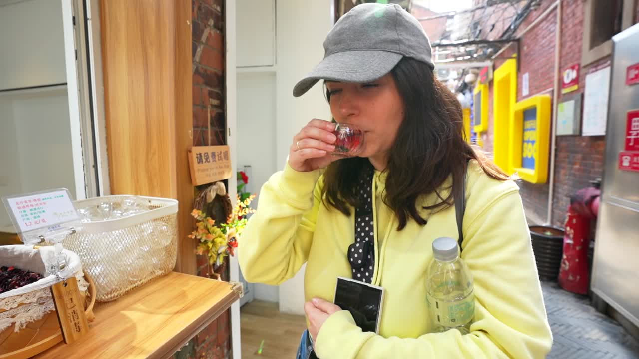 Tourist woman samples a small cup of red tea outside a Tianzifang shop in a lively lane. Shanghai, China