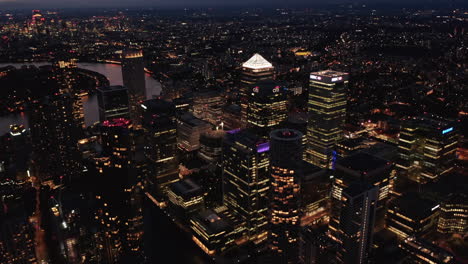 Deslizamiento-Aéreo-E-Inclinación-De-Edificios-De-Oficinas-Modernos-En-El-Centro-De-Negocios-De-Canary-Wharf.-Rascacielos-Iluminados-Por-La-Noche.-Londres,-Reino-Unido