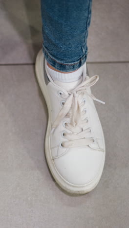 primer plano de zapatillas de deporte blancas de lona y pantalones de jean de mujer colocados en un piso limpio de azulejos en un centro comercial tocando su pie derecho, mostrando zapatos cómodos