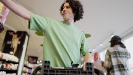 bottom view of a confident brunette guy with curly hair in a green t-shirt carries a cart and puts the goods he needs into it in a supermarket