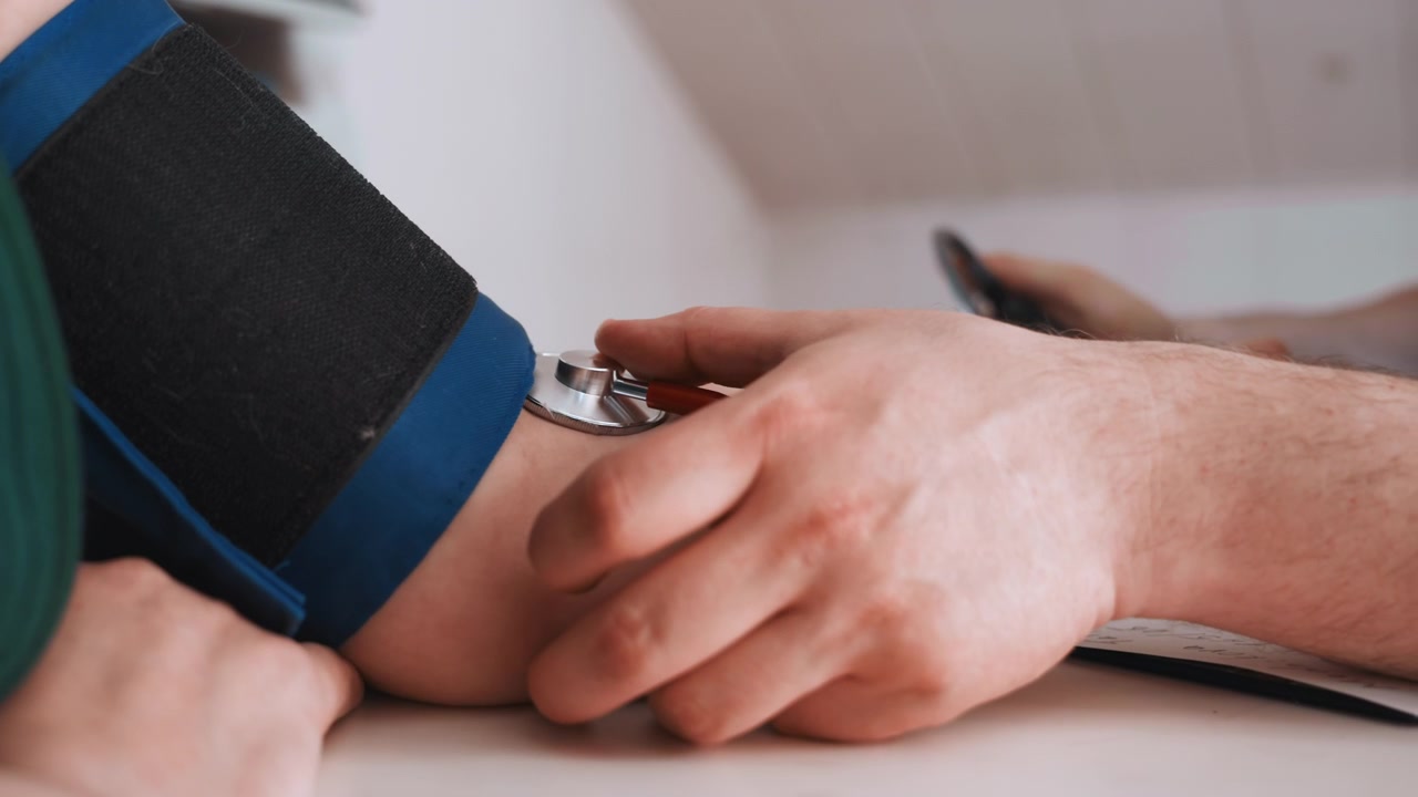 Bracciale per la pressione sul tavolo durante un controllo di telemedicina, l'infermiera regola il connettore mentre il paziente è seduto nelle vicinanze, laptop e appunti visibili, scena domestica tranquilla che enfatizza il monitoraggio e la consultazione a distanza