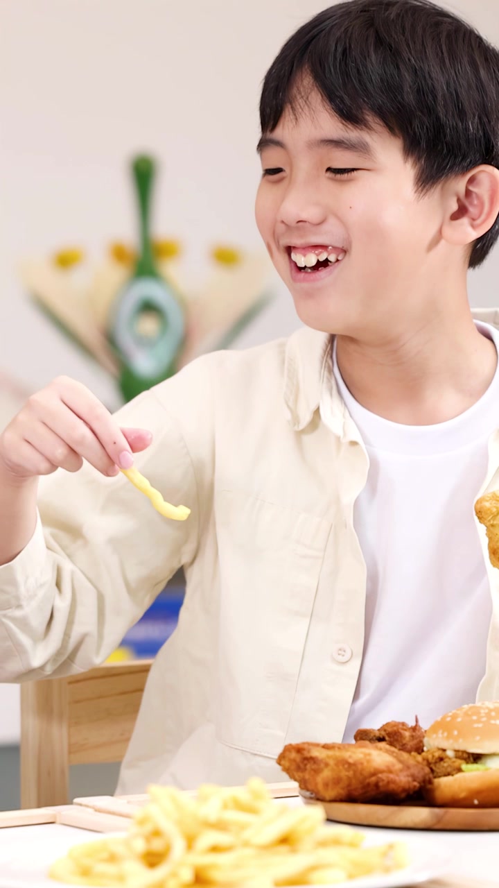 明るい教室のランチテーブルでフライドポテトとフライドチキンを楽しむ笑顔の男子生徒、ミディアムクローズアップ。