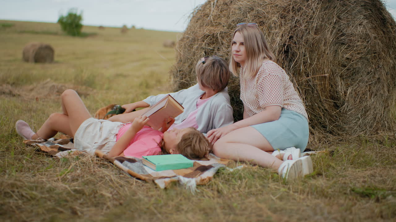 маленькая девочка в розовой рубашке читает свою книгу с зеленой обложкой, лежит на пикниковом одеяле с семьей, отдыхающей поблизости, мать сидит рядом, другая женщина отдыхает с собакой на поводке, глядя в расстояние