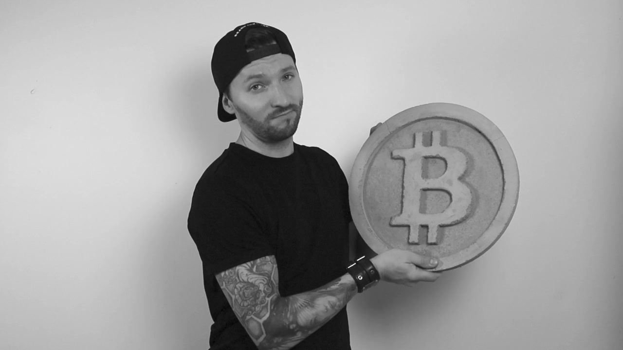 Gråtoneoptagelse af en tatoveret mand i sort tøj, der løfter en betonmodel af en bitcoin-mønt i slowmotion, mens han er glad og nikker godkendende foran en hvid baggrund