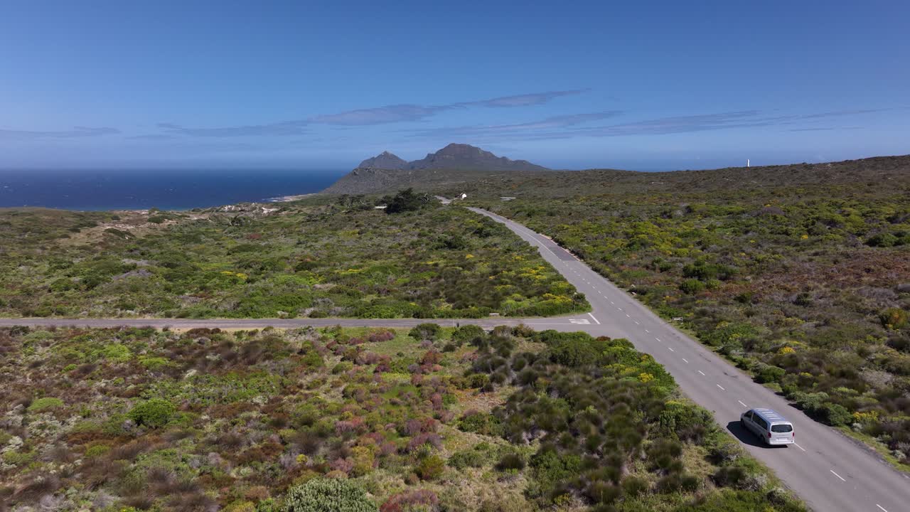푸른 덤불과 먼 언덕이 있는 밝고 맑은 날 해변 고속도로에서 운전하는 밴의 넓은 공중 샷