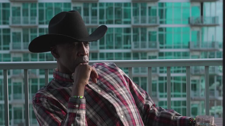 Uomo di colore con cappello da cowboy e un bicchiere di vetro, seduto su un balcone.