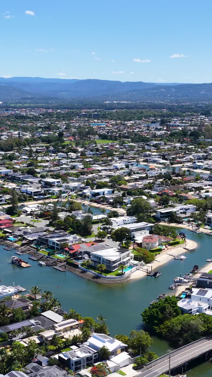 Un dron sobrevuela casas junto al canal, con cielos despejados y luz diurna brillante, revelando el paisaje suburbano de Gold Coast.