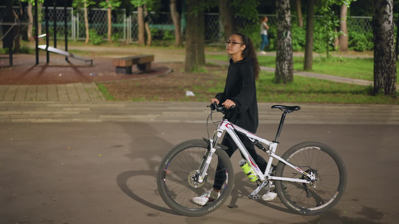 Urban Evening Stroll, Pedestrian Woman In Hoodie Riding Bike Along Shaded Pathway At Dusk, Calm Urban Environment With Woman Dressed Casually Biking Past Grove Of Trees During Soft Evening Glow