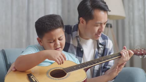 close up of asian father is teaching his son singing and playing the guitar on sofa at home