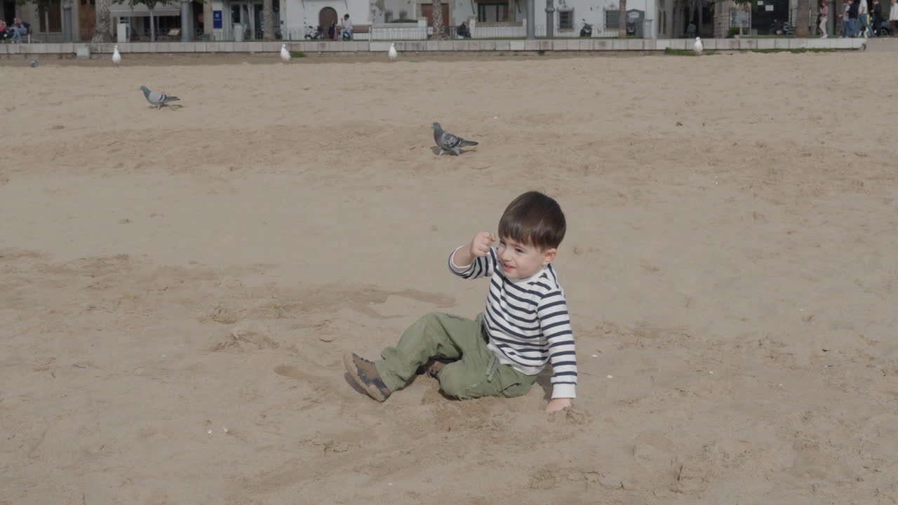 Toddler laughing and pointing while sitting in beach sand surrounded by pigeons walking and flying over his head