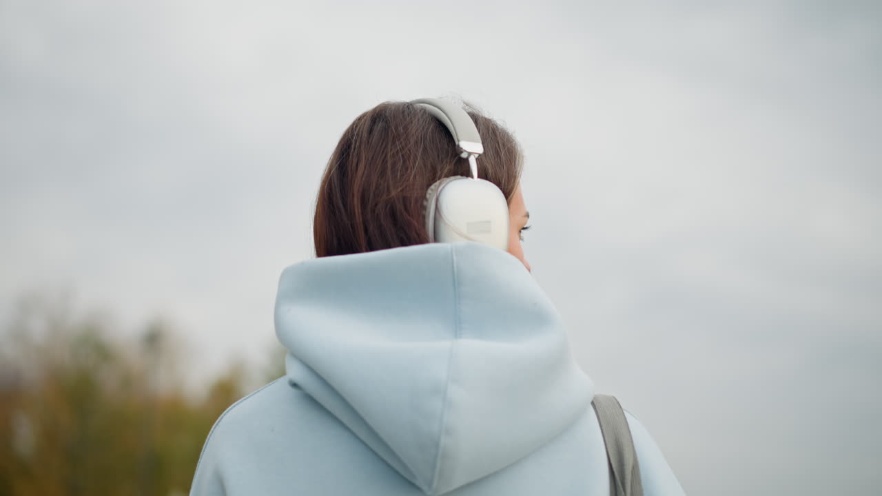 Back view of young lady in blue sweater and headphones walking in cool environment, blurred withered tree in background, adding a calm and serene vibe to the outdoor scene