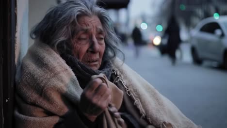 an elderly woman sits on the cold pavement, wrapped in a blanket, clearly distressed by the chilly evening air. the surrounding city lights and passersby create a stark contrast to her situation.