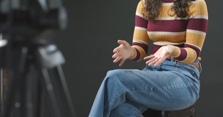 Camera starting recording, woman uncrossing legs and gesturing palms open while speaking on stool