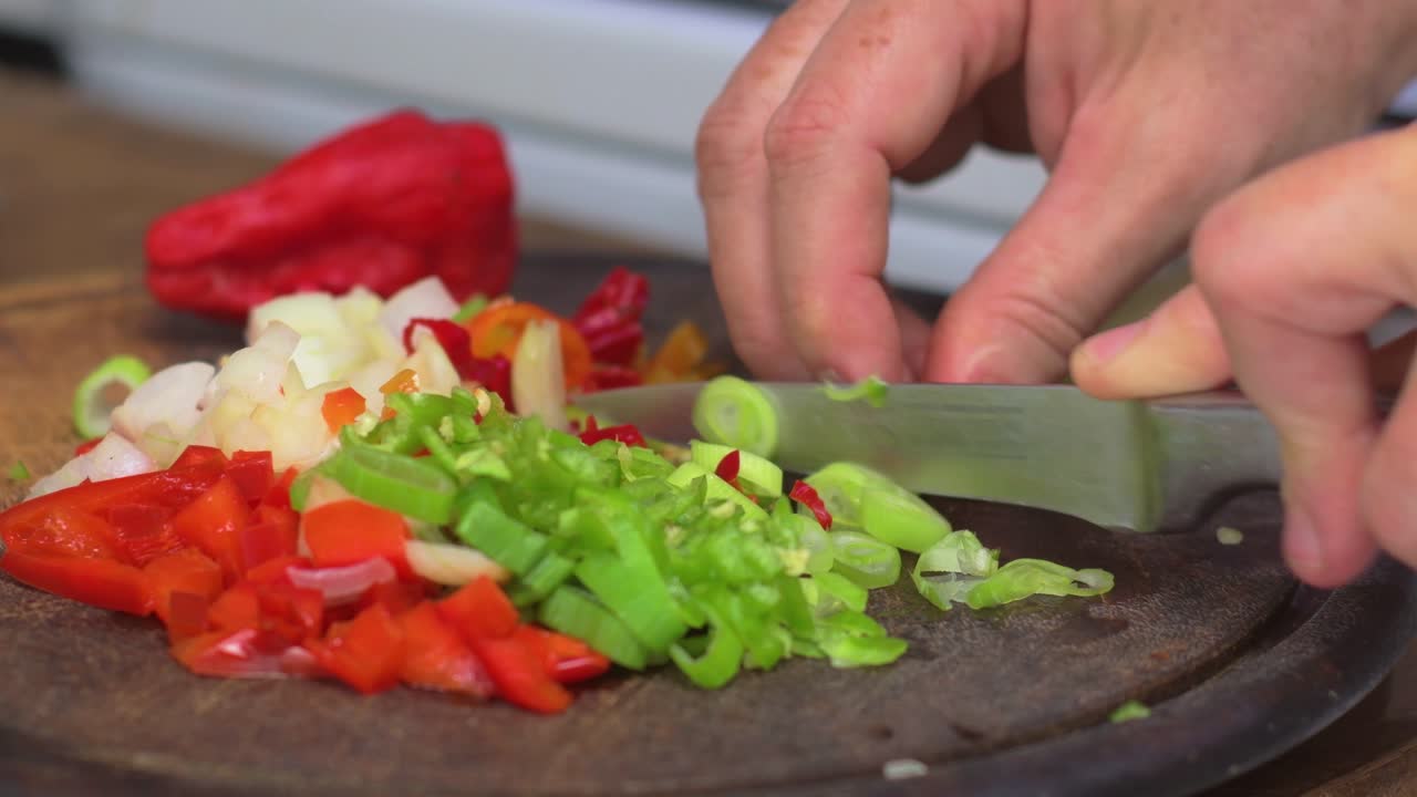Close up of hands cutting scallion, pepper, and onion on wooden board in kitchen