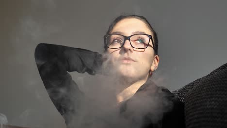 young woman releasing cigarette smoke through nose while sitting in room looking on sunlight coming from window, slow motion