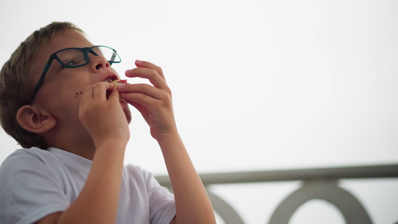se ve a un niño sosteniendo un rollo de salchichas, observándolo cuidadosamente antes de ponerlo en su boca, mira hacia la derecha con una sonrisa mientras disfruta de su bocadillo