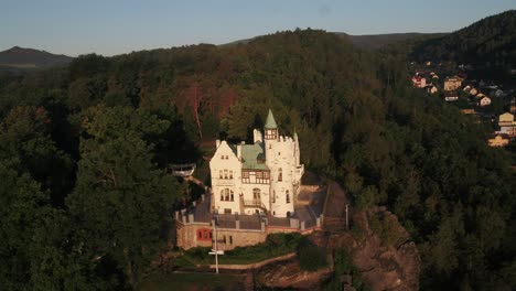 drone rises slowly behind trees to reveal decin castle in the distance, bathed in early morning sunlight