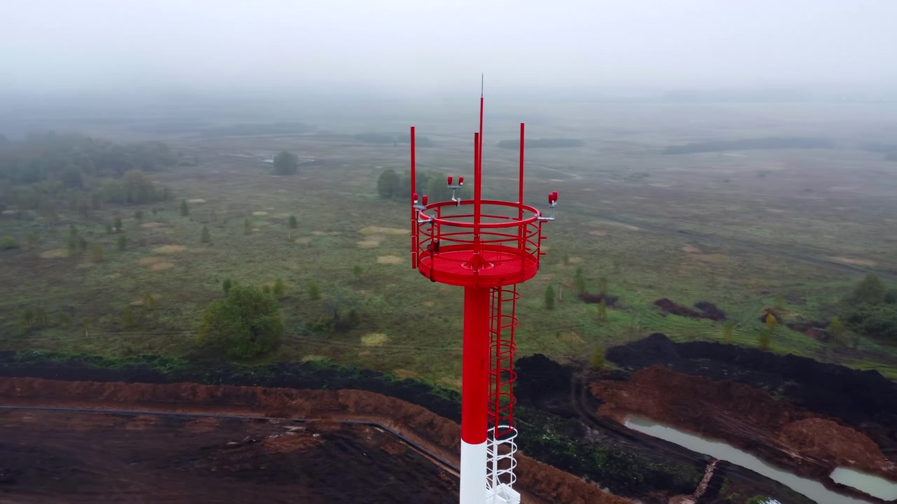 Una torre di comunicazione rossa e bianca si erge su un'altura sopra un ampio paesaggio rurale, vista da un'angolazione aerea elevata. La nebbia ammorbidisce l'orizzonte mentre