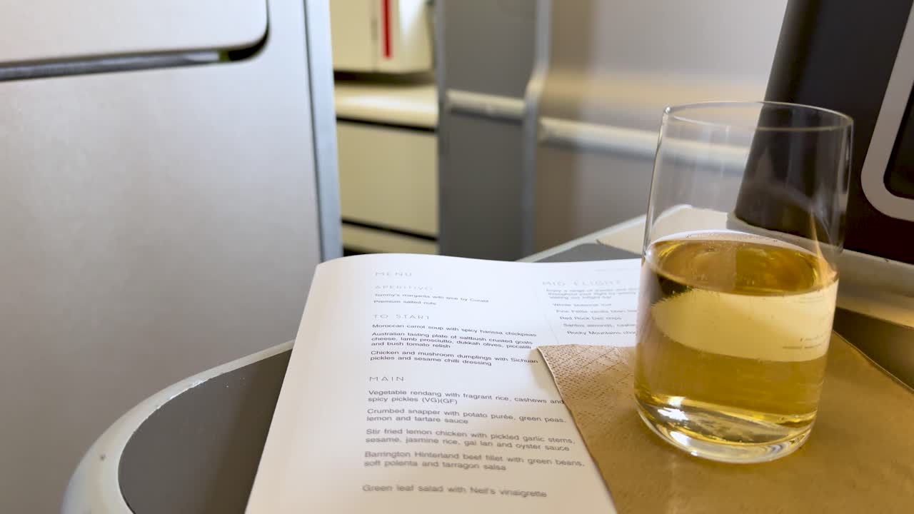 Glass of sparkling beverage on airplane tray table beside printed menu, steady camera, soft lighting