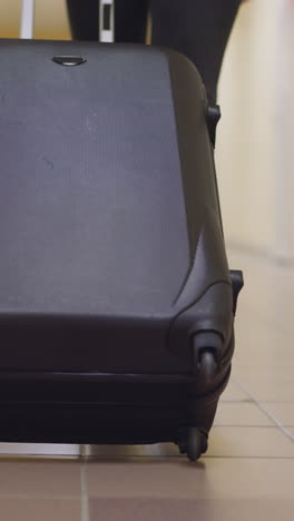 woman carries big black suitcase on tile floor of corridor of hotel after arrival. female tourist takes baggage to personal room close backside view