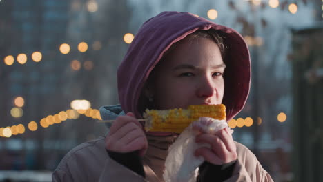 mujer joven disfrutando de maíz fresco en un palo envuelto en una bolsa blanca con vapor que se eleva en un entorno al aire libre, luces bokeh en un fondo borroso, bocadillo de clima frío en la ciudad