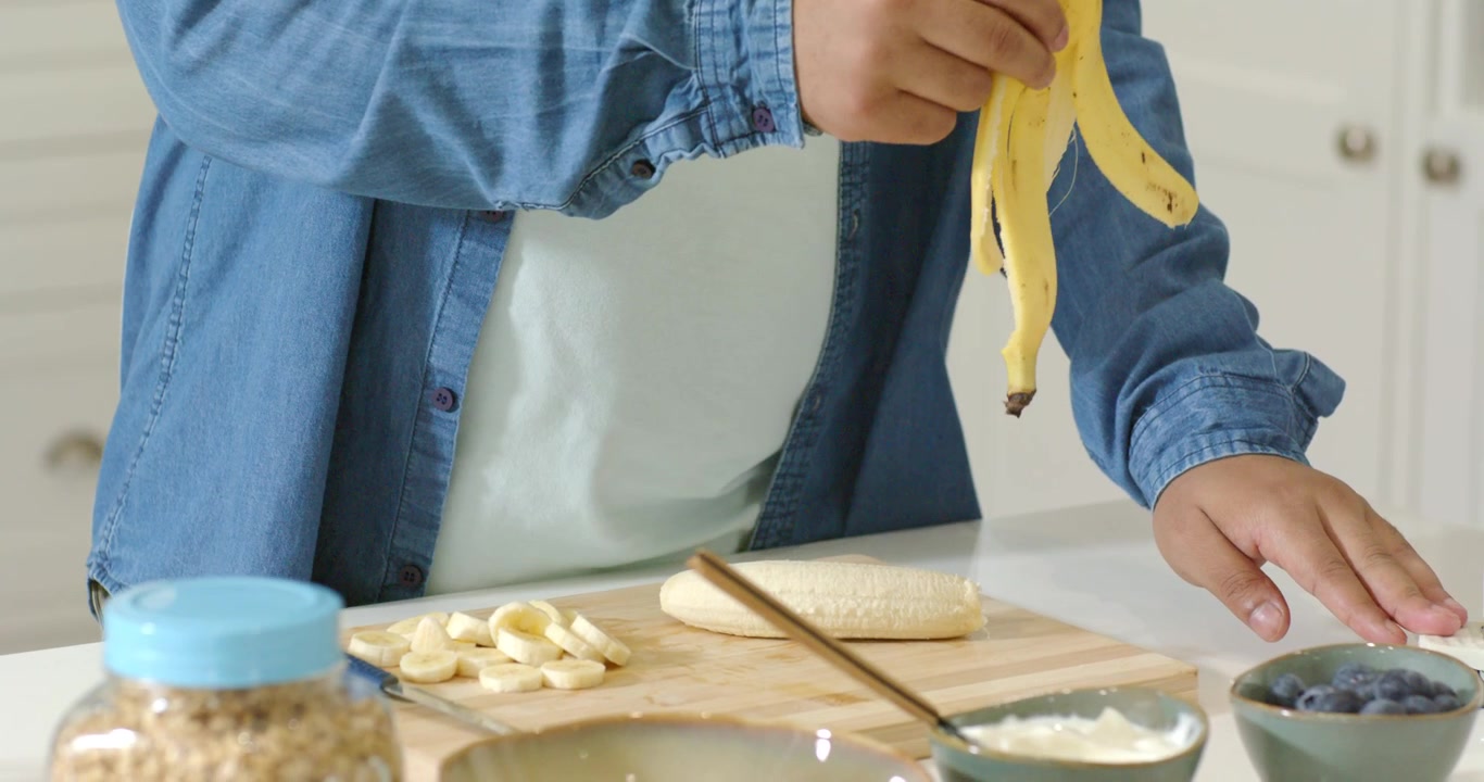 Hombre pelando un plátano en la encimera de la cocina, colocándolo en una tabla, tirando la cáscara en un cubo blanco para el desayuno