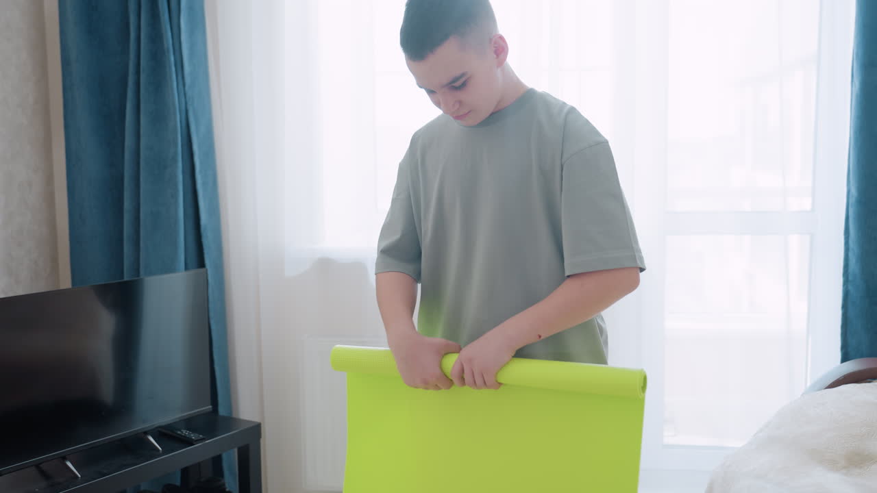 Young boy folds bright green fitness exercise mat inside living room, preparing space for workout, casual lifestyle scene showing health awareness