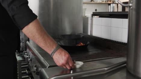 caucasian male chef frying vegetables in frying pan with bursting fire in kitchen, slow motion