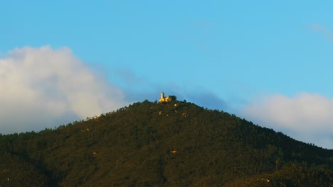 cinematic, serene view of lone building atop forest-covered mountain with blue skies in varazze, italy