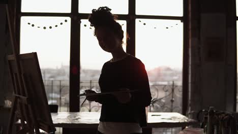 young sweet female artist dancing while drawing her picture with paint palette in her hands, in the loft styled art studio.