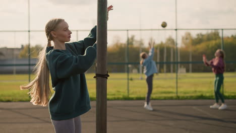 side view of woman holding volleyball net while two other women are playing volleyball in the background, outdoor sports setting with clear sky and distant action