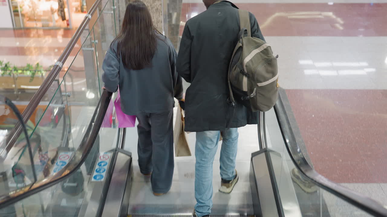 Rear view of siblings riding down escalator inside bright spacious mall, holding shopping bags and engaging in conversation while glass railings reflect light and palm tree decor