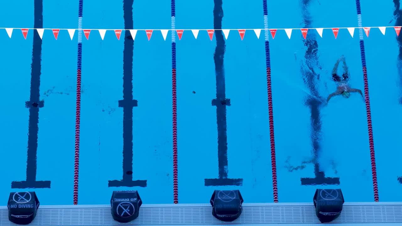 A swimmer practices freestyle strokes in a clear blue pool, captured from above in bright daylight