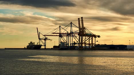 timelapse small tug boat guides a huge container ship into a sea port while another moored ship is being unloaded by container cranes at sunset in the port of malaga in the costa del sol, spain