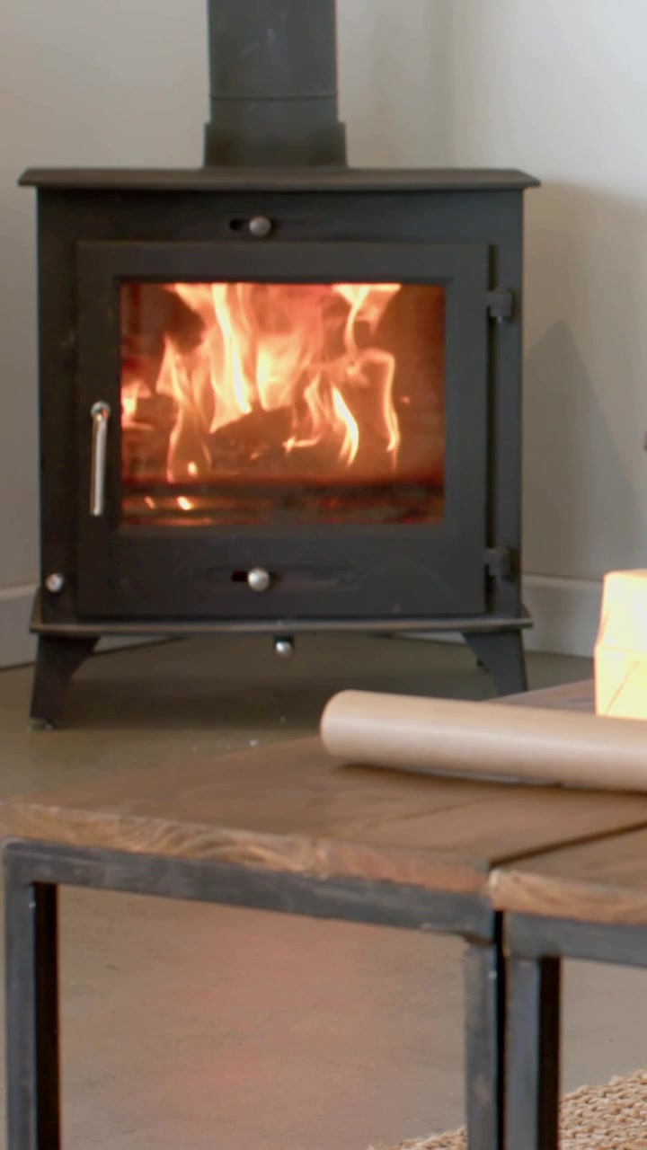 Video verticale: Stufa a legna nera che arde con un ceppo dietro il vetro in un angolo di casa, la telecamera si sposta lentamente.
