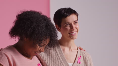 retrato de estudio de dos mujeres que llevan cintas rosadas de concienciación sobre el cáncer de mama abrazándose contra un fondo rosa