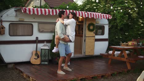 Happy-young-lovely-couple-having-fun-near-trailer-in-the-park,-girl-jumping-on-man's-arms,-he's-whirling-her-around.-Retro---american-exterior